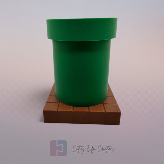 A 3D-printed pen holder designed to resemble a green pipe from a classic video game, sitting on a brown, block-patterned base. The cylindrical holder is vibrant green with a slightly wider rim at the top. The brown base mimics the look of pixelated bricks.