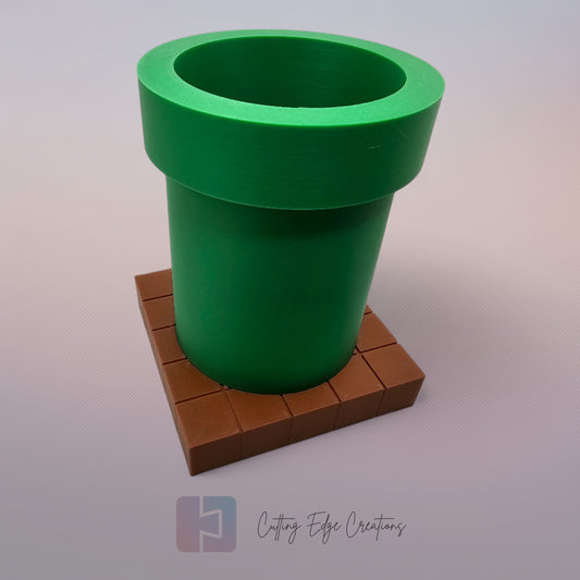 A 3D-printed pen holder designed to resemble a green pipe from a classic video game, sitting on a brown, block-patterned base. The cylindrical holder is vibrant green with a slightly wider rim at the top. The brown base mimics the look of pixelated bricks.