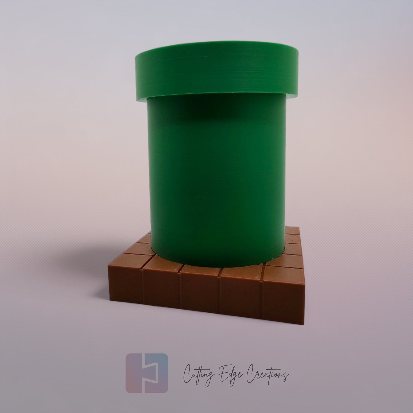 A 3D-printed pen holder designed to resemble a green pipe from a classic video game, sitting on a brown, block-patterned base. The cylindrical holder is vibrant green with a slightly wider rim at the top. The brown base mimics the look of pixelated bricks.