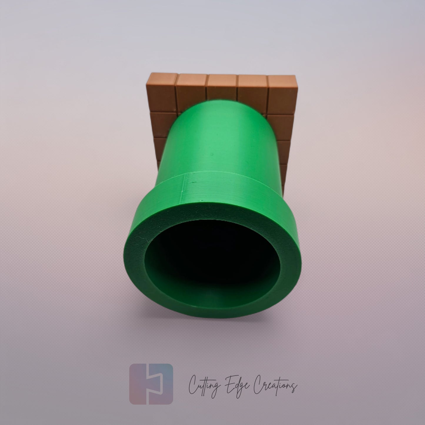 A 3D-printed pen holder designed to resemble a green pipe from a classic video game, sitting on a brown, block-patterned base. The cylindrical holder is vibrant green with a slightly wider rim at the top. The brown base mimics the look of pixelated bricks.