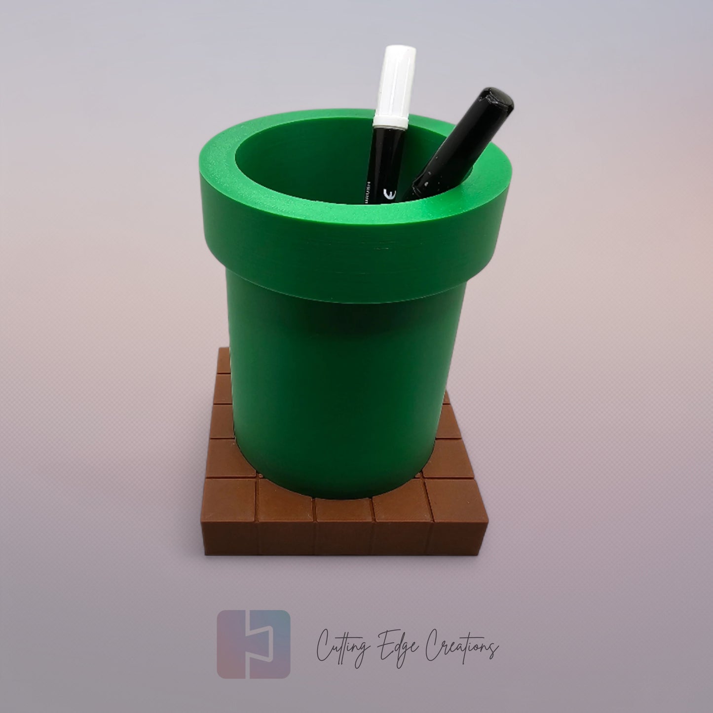 A 3D-printed pen holder designed to resemble a green pipe from a classic video game, sitting on a brown, block-patterned base. The cylindrical holder is vibrant green with a slightly wider rim at the top. The brown base mimics the look of pixelated bricks.