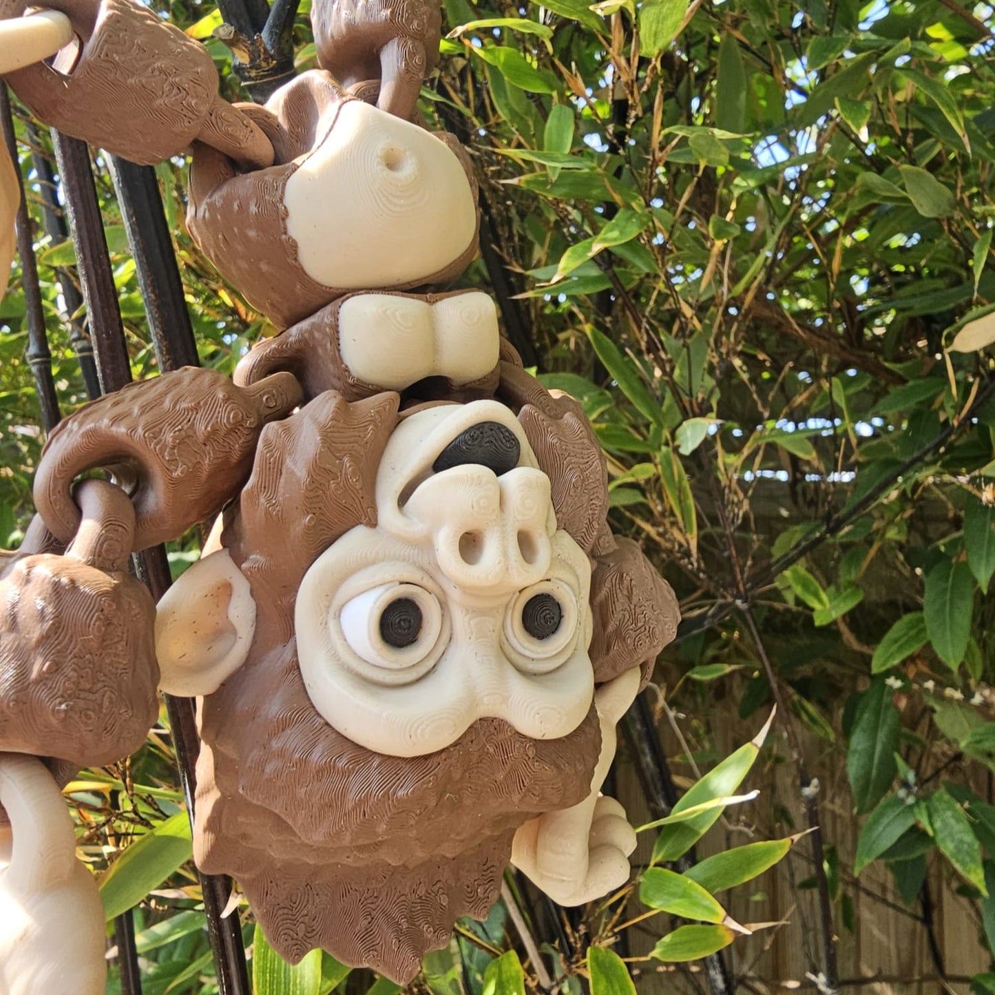 3D printed articulated Mega Gorilla toy hanging upside down from black poles in a garden, with hook-shaped hands and feet, surrounded by lush green bamboo leaves.