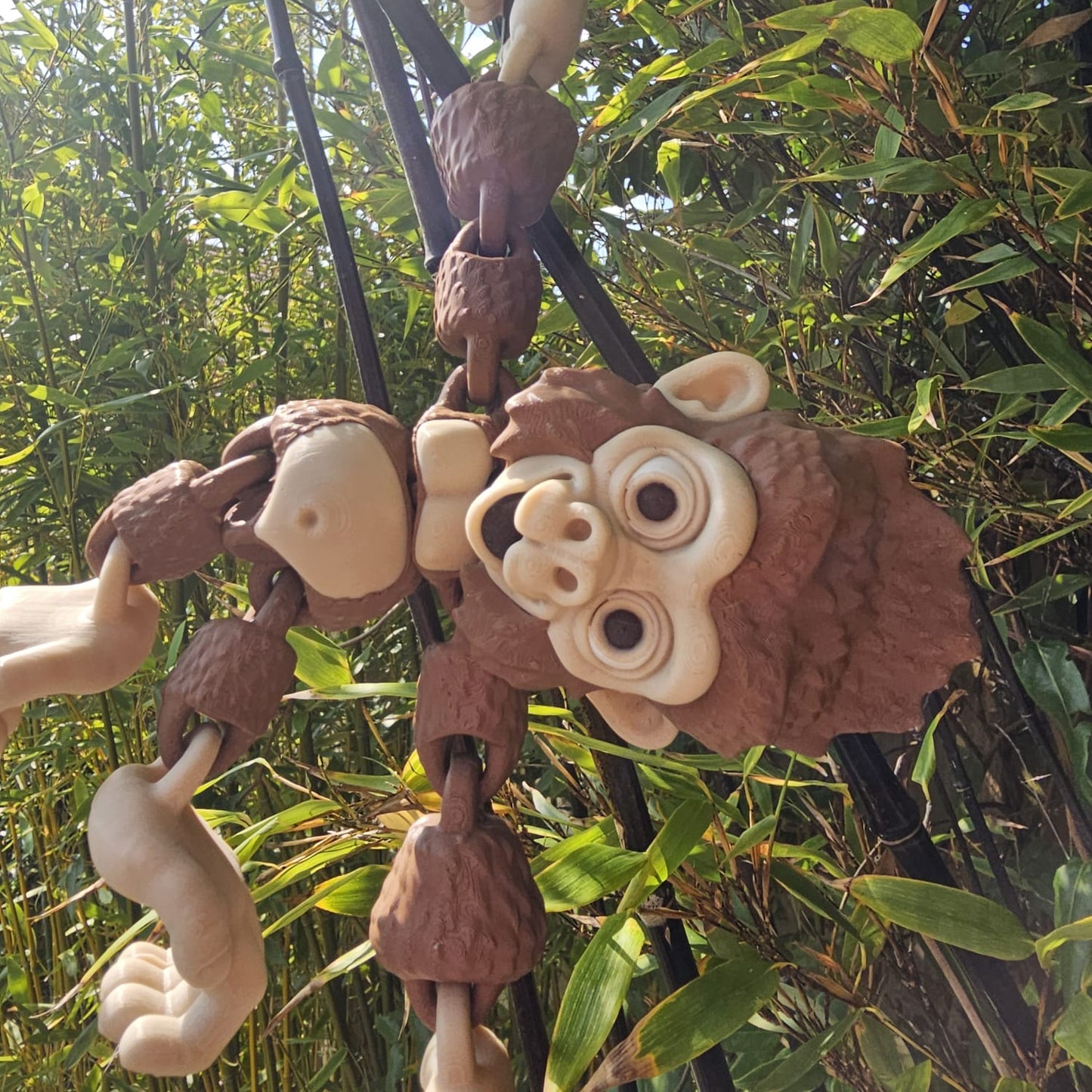 3D printed articulated Mega Gorilla toy hanging upside down from black poles in a garden, with hook-shaped hands and feet, surrounded by lush green bamboo leaves.