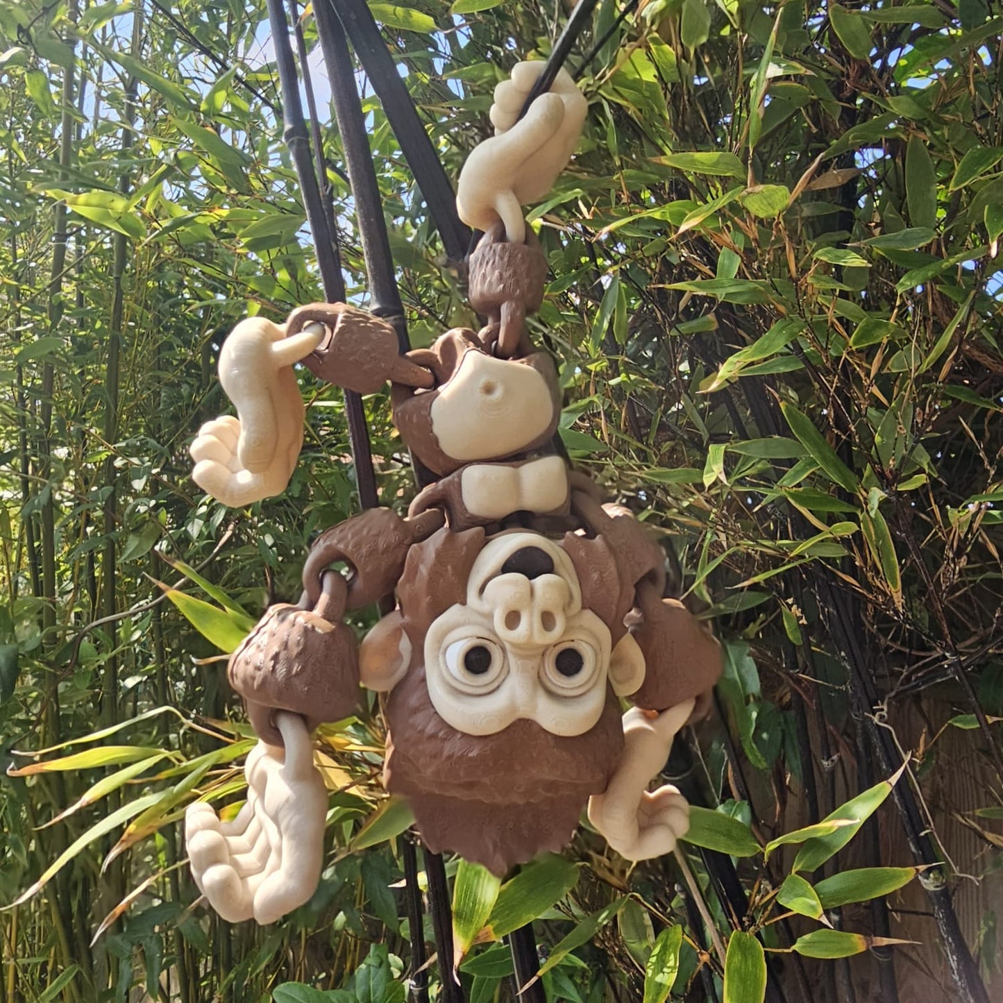 3D printed articulated Mega Gorilla toy hanging upside down from black poles in a garden, with hook-shaped hands and feet, surrounded by lush green bamboo leaves.