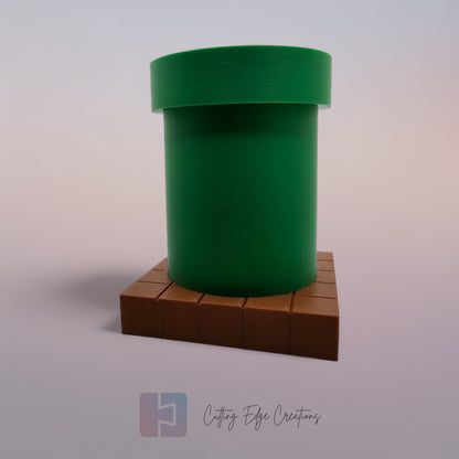A 3D-printed pen holder designed to resemble a green pipe from a classic video game, sitting on a brown, block-patterned base. The cylindrical holder is vibrant green with a slightly wider rim at the top. The brown base mimics the look of pixelated bricks.