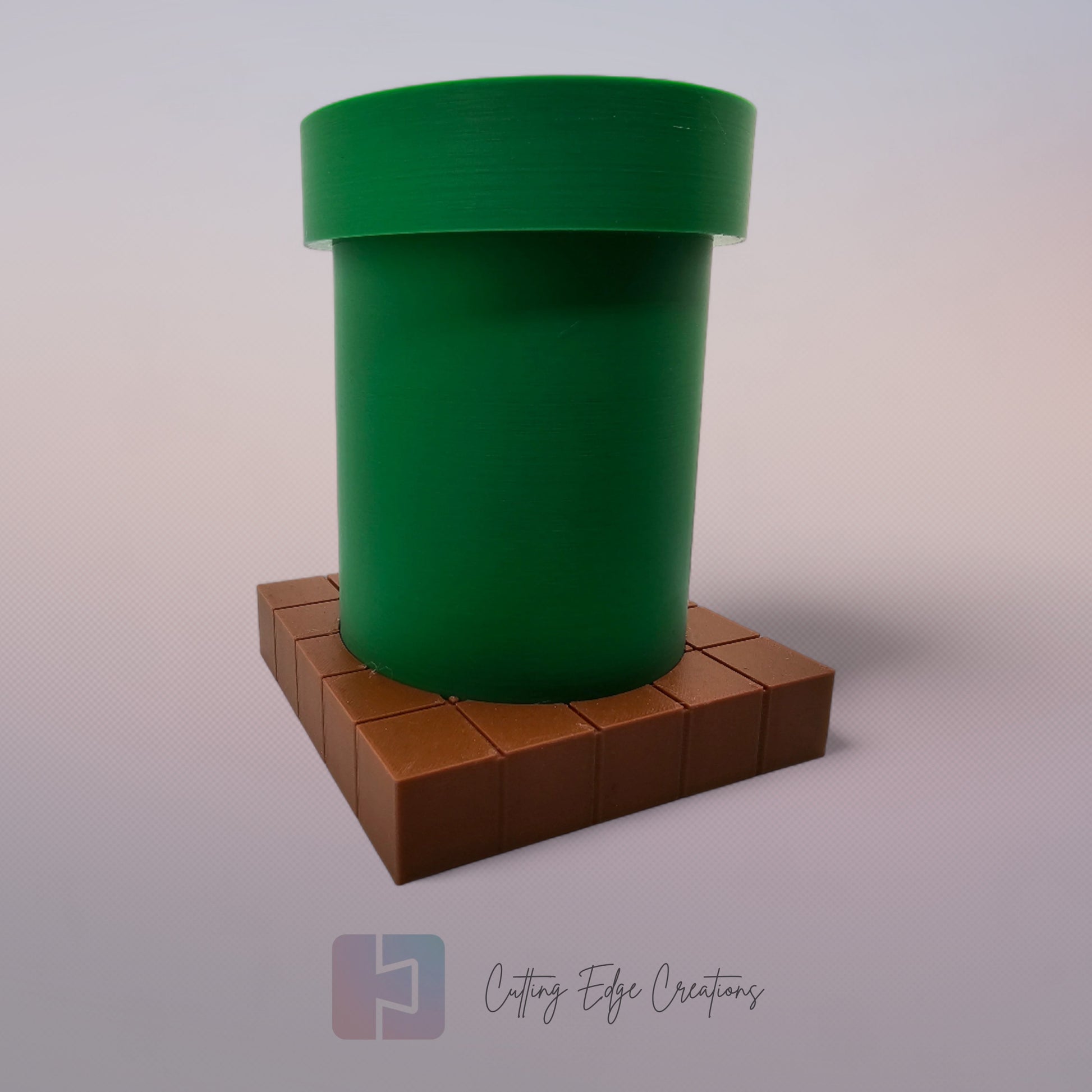 A 3D-printed pen holder designed to resemble a green pipe from a classic video game, sitting on a brown, block-patterned base. The cylindrical holder is vibrant green with a slightly wider rim at the top. The brown base mimics the look of pixelated bricks.