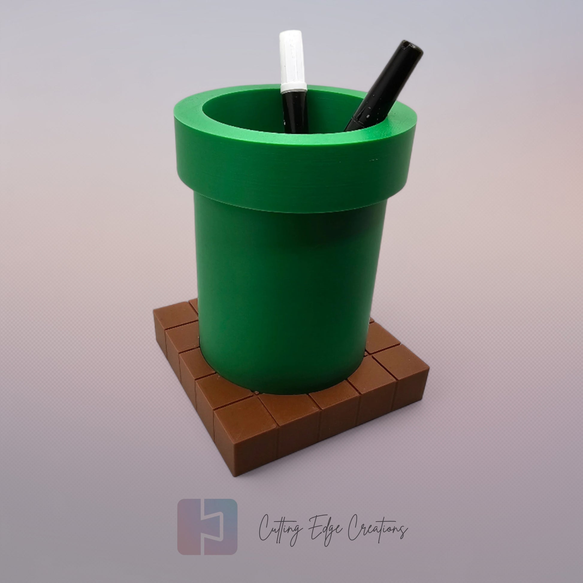 A 3D-printed pen holder designed to resemble a green pipe from a classic video game, sitting on a brown, block-patterned base. The cylindrical holder is vibrant green with a slightly wider rim at the top. The brown base mimics the look of pixelated bricks.