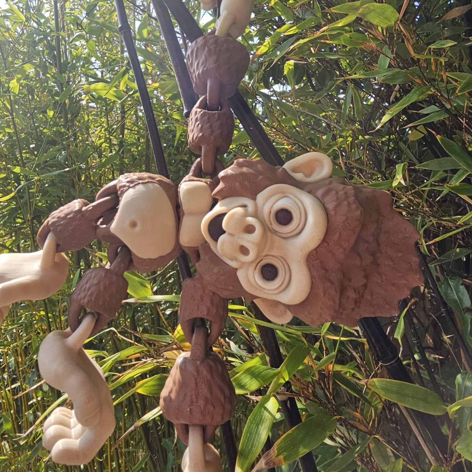 3D printed articulated Mega Gorilla toy hanging upside down from black poles in a garden, with hook-shaped hands and feet, surrounded by lush green bamboo leaves.