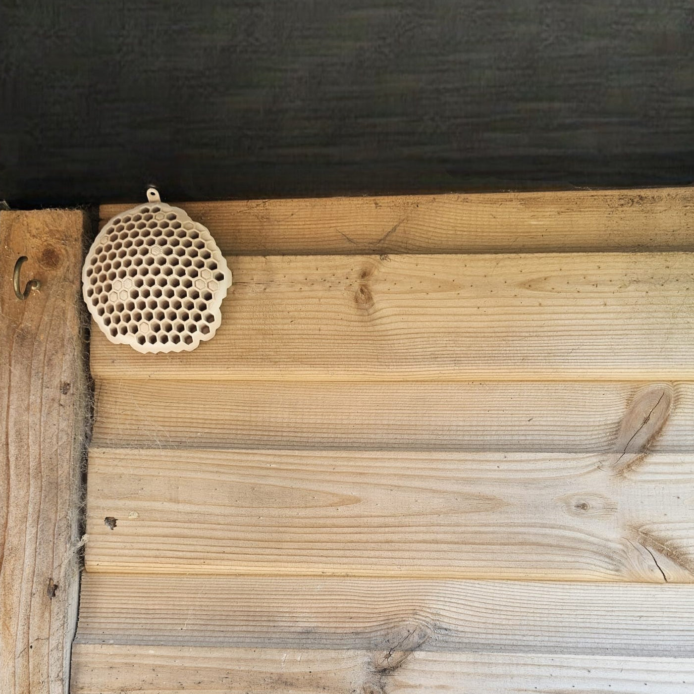 A 3D printed fake wasp nest with a honeycomb pattern, mounted under a wooden shed roof to deter real wasps.